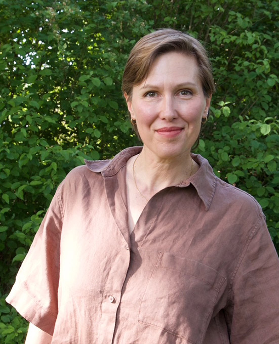 Eine Person mit kurzen braunen Haaren, Katja Karle, steht im Freien vor grünem Laub, trägt ein locker sitzendes braunes Hemd und lächelt leicht in die Kamera.